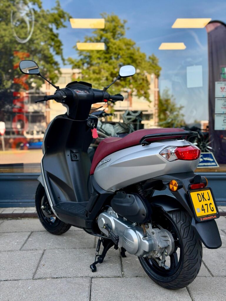 Zilveren scooter met rood zadel geparkeerd voor een winkelraam op een zonnige dag.