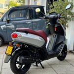 Zilverkleurige scooter met rood zadel geparkeerd voor een garage, grijze microcar erachter.