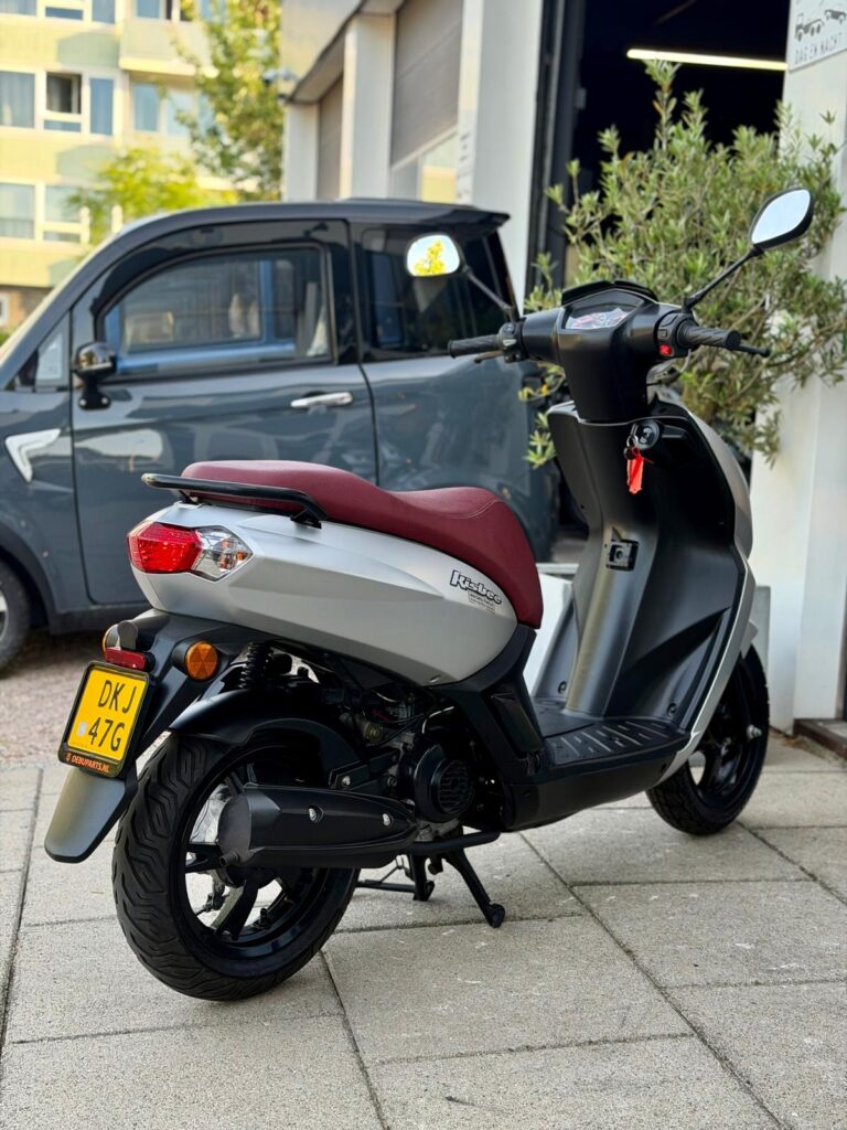 Zilverkleurige scooter met rood zadel geparkeerd voor een garage, grijze microcar erachter.