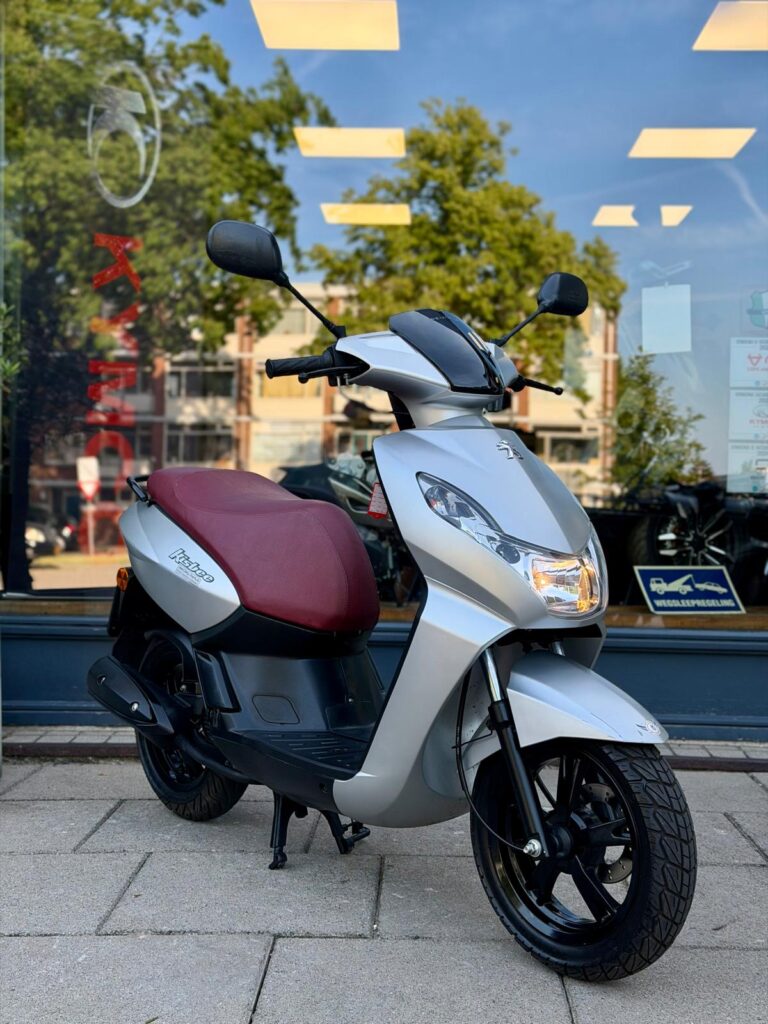 Zilveren scooter met bordeauxrood zadel, geparkeerd voor een winkelruit met weerspiegelde bomen.