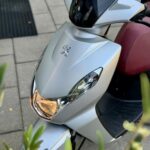 Close-up van een zilvergrijze Peugeot-scooter met rood zadel, koplamp aan, geparkeerd op stoep tussen groen.
