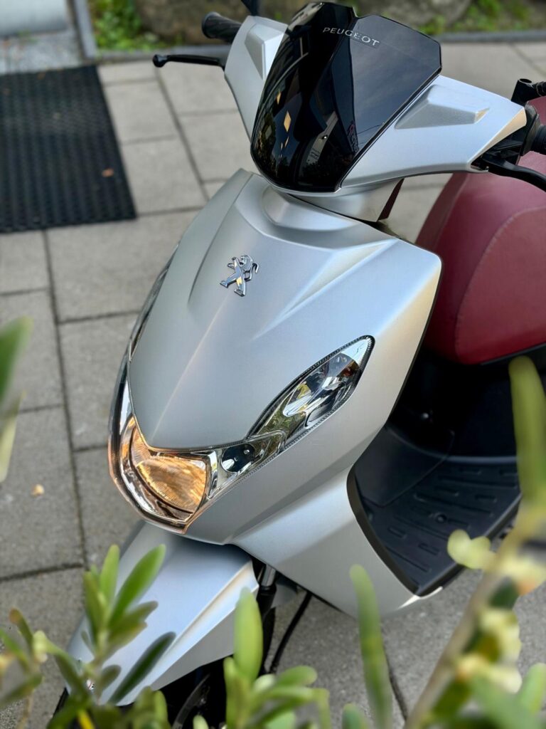 Close-up van een zilvergrijze Peugeot-scooter met rood zadel, koplamp aan, geparkeerd op stoep tussen groen.