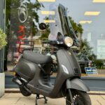 Mat zwarte scooter met hoog windscherm op standaard, geparkeerd voor een scooterwinkel