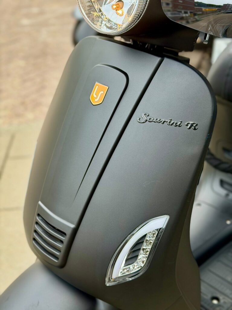 Detail van de voorkap van een matte grijze scooter met koplamp, LED-knipperlicht en Sourini R-embleem.