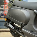Close-up van een matte zwarte La Souris-scooter met uitlaat, achterwiel en bagagerek.