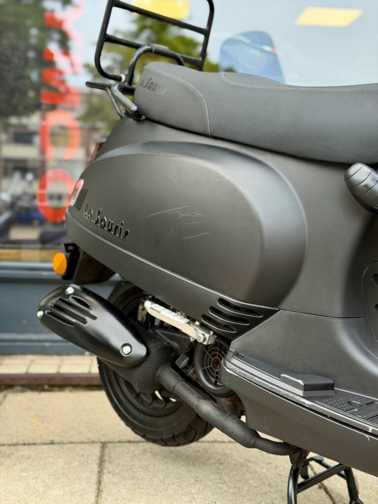 Close-up van een matte zwarte La Souris-scooter met uitlaat, achterwiel en bagagerek.