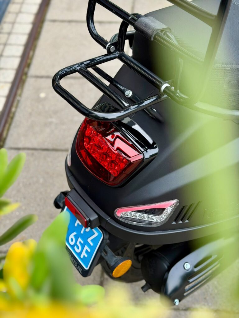 Close-up van de achterkant van een zwarte scooter met bagagedrager, remlicht en blauwe kentekenplaat.