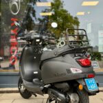 Matzwarte La Souris-scooter met bagagedrager en blauw kenteken, geparkeerd voor een winkelraam.