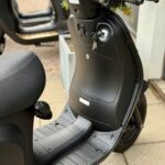 Close-up van een zwarte scooter met sleutels in het contact, geparkeerd op de stoep.