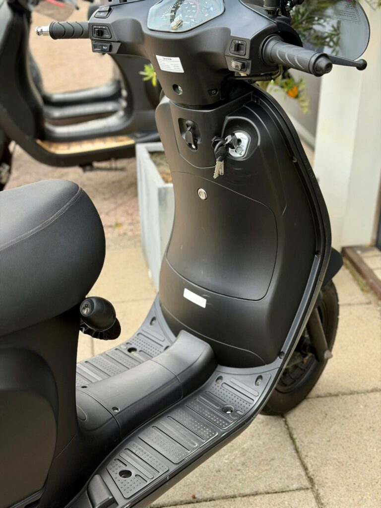 Close-up van een zwarte scooter met sleutels in het contact, geparkeerd op de stoep.