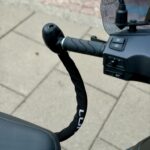 Scooterstuur met spiegel en zwarte ketting als stuurslot, bevestigd tussen handvat en zadel.