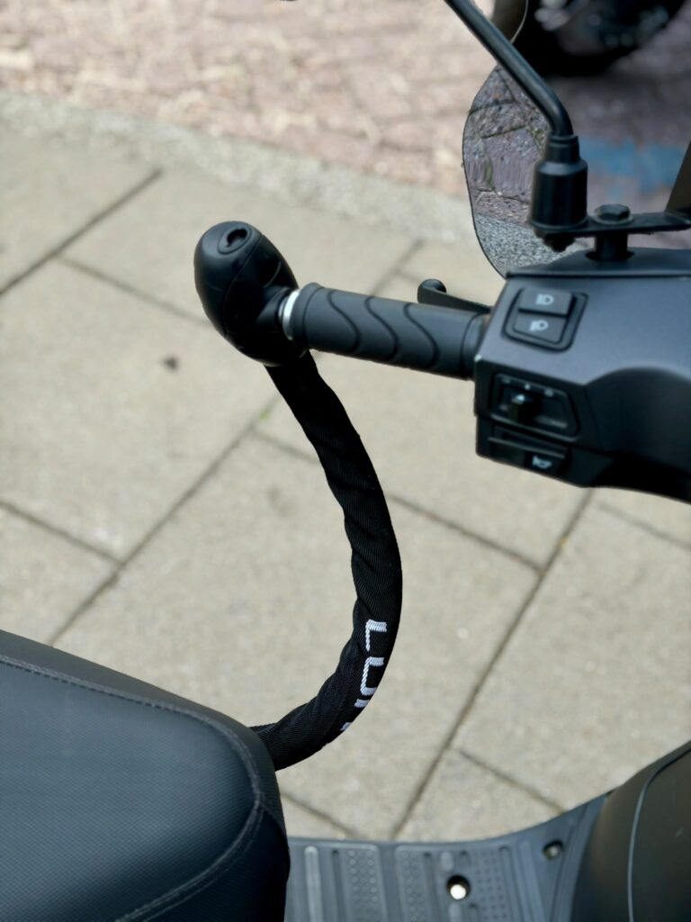 Scooterstuur met spiegel en zwarte ketting als stuurslot, bevestigd tussen handvat en zadel.