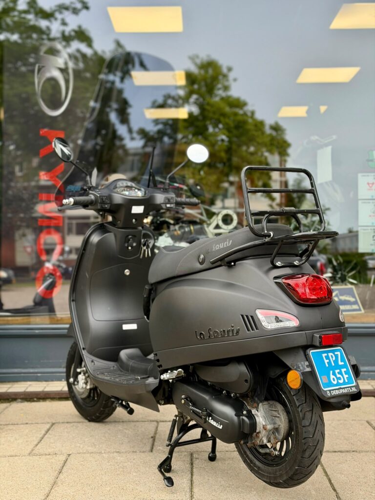 Matzwarte La Souris-scooter met bagagedrager en blauw kenteken, geparkeerd voor een winkelraam.