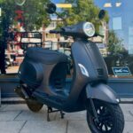 Matte zwarte scooter geparkeerd op de stoep voor een winkel met glazen pui