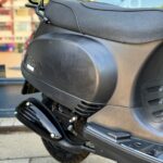 Detail van de rechterzijkant van een zwarte scooter met uitlaat, zadel en bagagedrager.