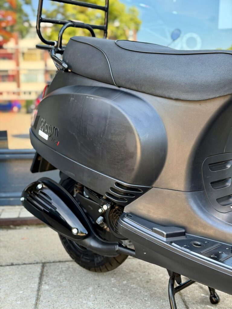Detail van de rechterzijkant van een zwarte scooter met uitlaat, zadel en bagagedrager.