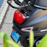 Achterkant van een zwarte scooter met bagagedrager, remlicht en blauw kenteken op de stoep.