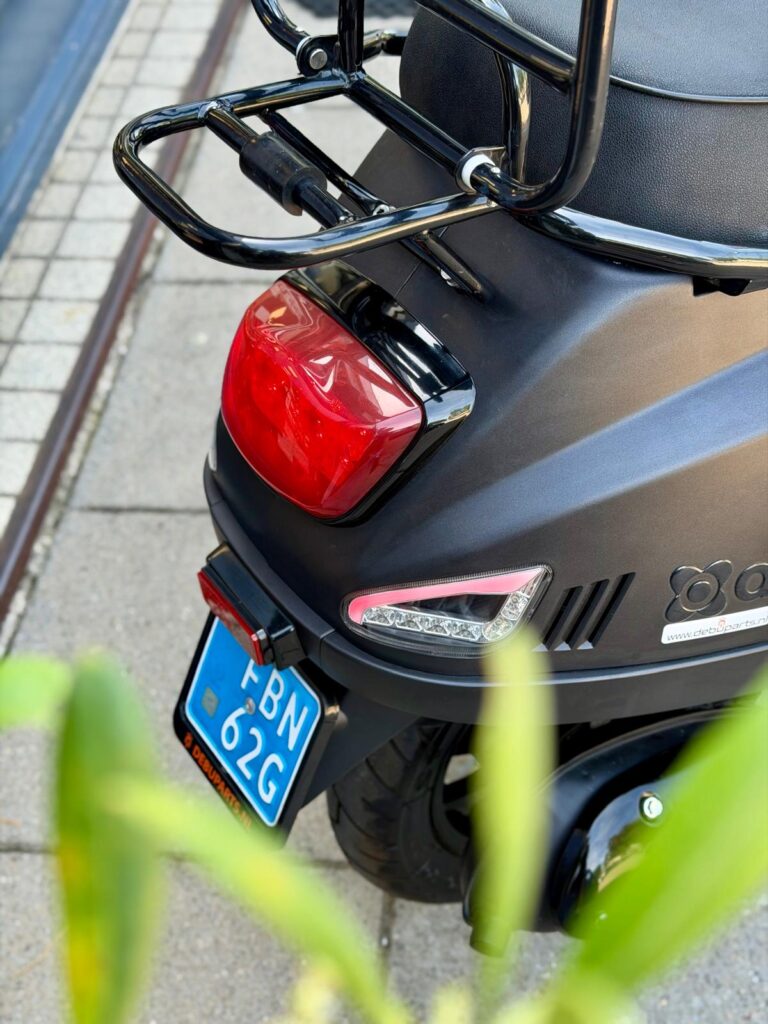 Achterkant van een zwarte scooter met bagagedrager, remlicht en blauw kenteken op de stoep.