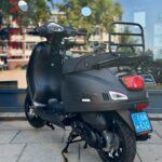 Mat zwarte scooter met blauw kenteken, geparkeerd op standaard voor een winkelpui.