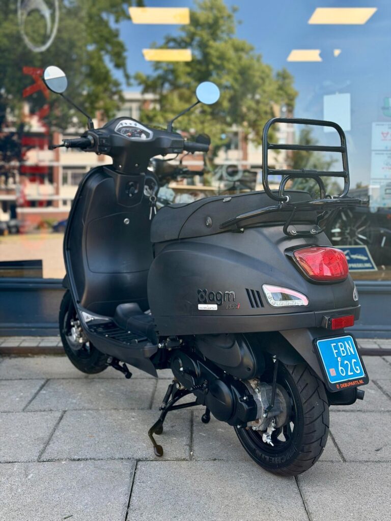Mat zwarte scooter met blauw kenteken, geparkeerd op standaard voor een winkelpui.
