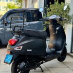 Matzwarte scooter met bagagedrager en blauwe kentekenplaat, geparkeerd voor een werkplaats.