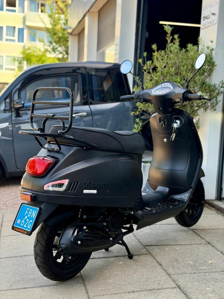 Matzwarte scooter met bagagedrager en blauwe kentekenplaat, geparkeerd voor een werkplaats.