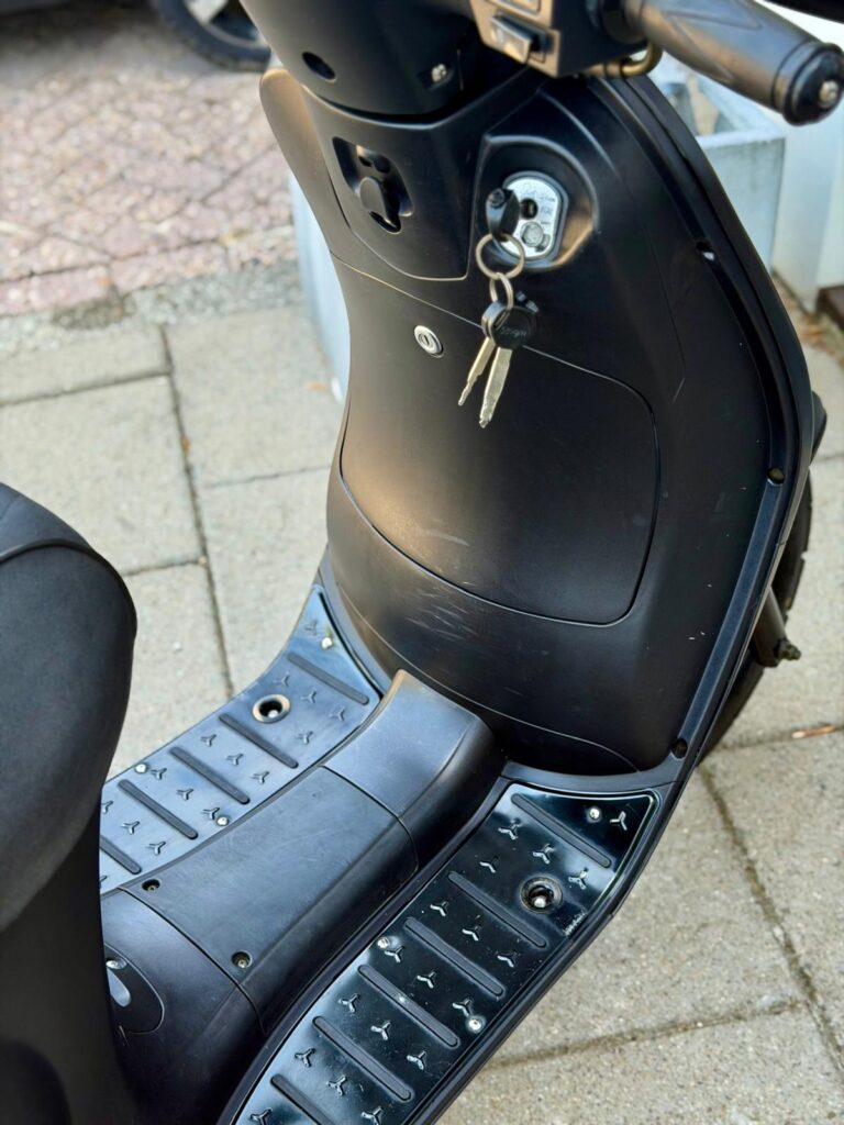 Close-up van een zwarte scooter met sleutels in het contact en de treeplank op straat.