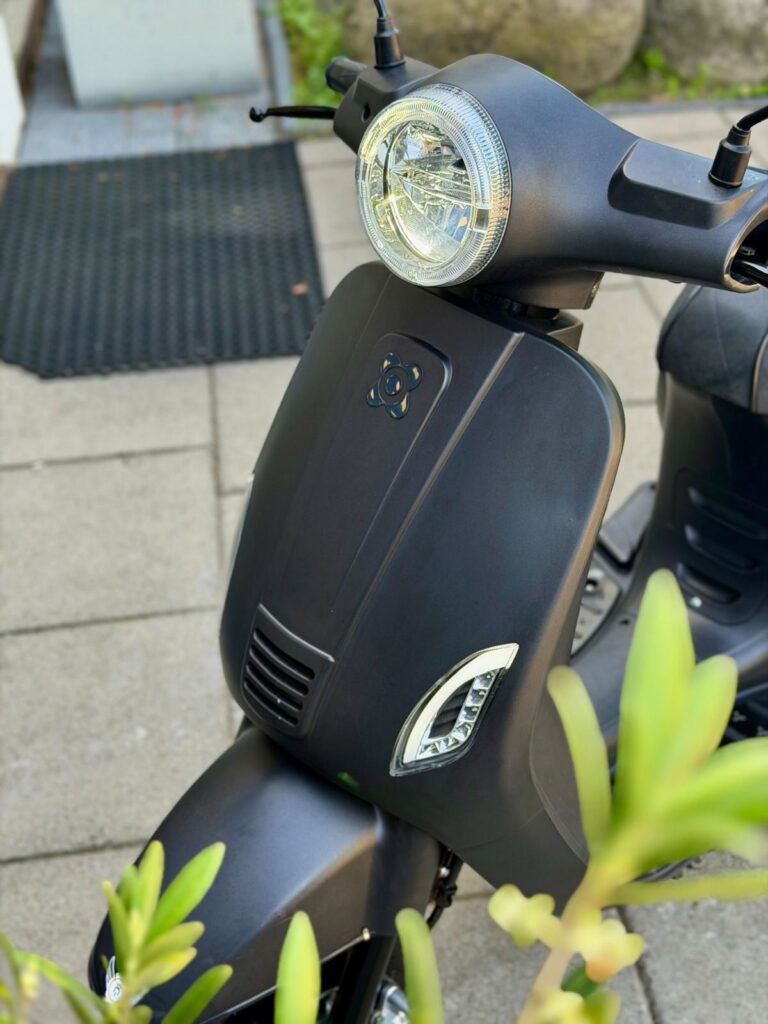 Close-up van de voorkant van een matte zwarte scooter met ronde koplamp en richtingaanwijzers.