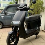 Matzwart elektrische scooter geparkeerd op trottoir voor gebouw en auto.