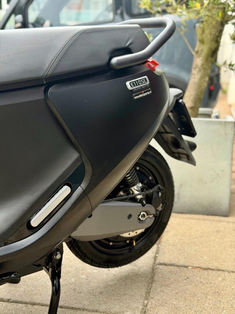 Close-up van de achterkant van een zwarte elektrische scooter met zadel, bagagedrager en achterwiel.