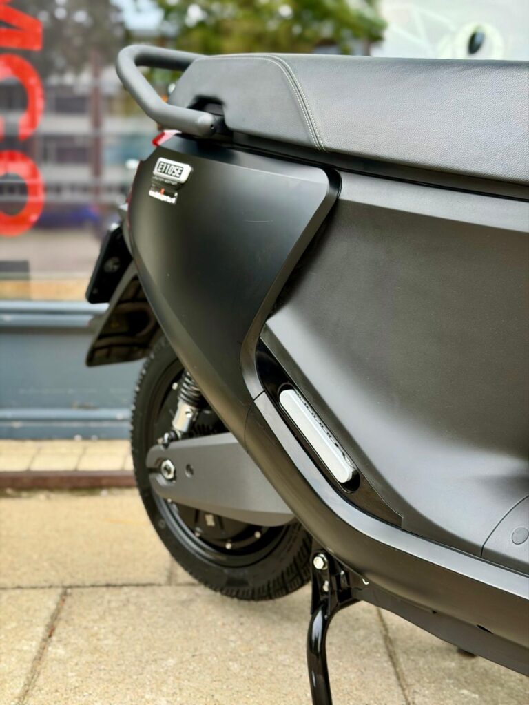 Close-up van de zijkant van een matzwarte scooter op de standaard, met zadel, handgreep en achterwiel.