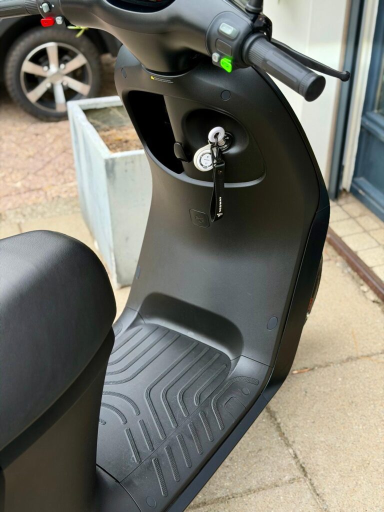 Close-up van een zwarte elektrische scooter met sleutel in het contact en rubberen voetplaat.