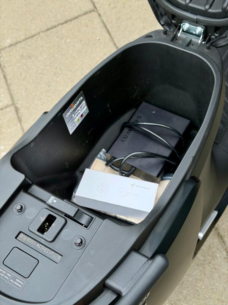 Open opbergruimte onder een scooterzadel met Segway-doosje en kabels.
