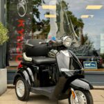 Zwarte driewielscooter met hoog windscherm, geparkeerd op de stoep voor een winkel.