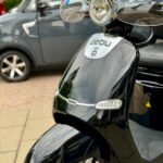 Close-up van een zwarte elektrische scooter met windscherm, geparkeerd op de stoep met auto op de achtergrond.