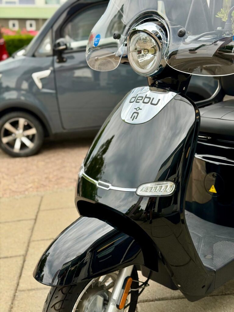 Close-up van een zwarte elektrische scooter met windscherm, geparkeerd op de stoep met auto op de achtergrond.