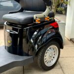 Close-up van een zwarte scootmobiel met zitting, armleuningen en achterwiel op het trottoir.