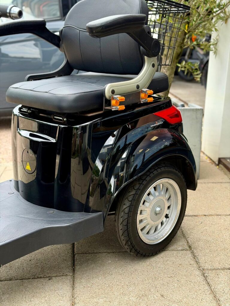 Close-up van een zwarte scootmobiel met zitting, armleuningen en achterwiel op het trottoir.