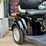 Close-up van de achterzijde van een zwarte scootmobiel met zitting, armleuningen en achterwiel op de stoep.
