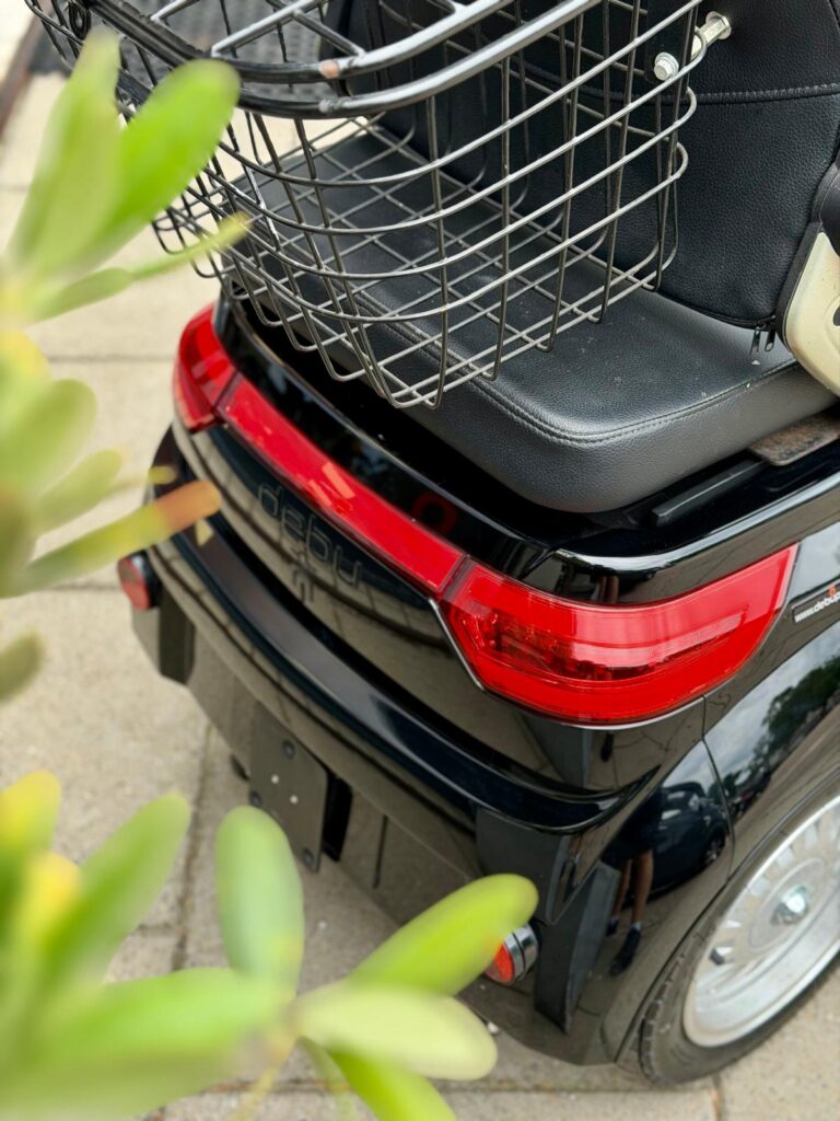 Achterkant van een zwarte scootmobiel met rood achterlicht en metalen bagagemand, plant op voorgrond.