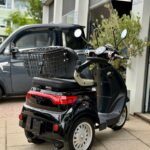 Zwarte driewielige scootmobiel met mandje, geparkeerd voor een garage.
