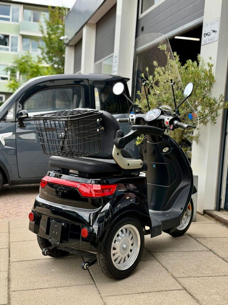 Zwarte driewielige scootmobiel met mandje, geparkeerd voor een garage.