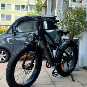 Zwarte fatbike e-bike met brede banden en koplamp, geparkeerd op stoep voor een gebouw met auto ernaast.