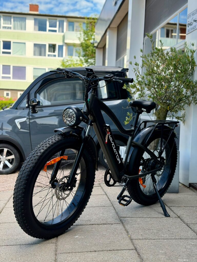 Zwarte fatbike e-bike met brede banden en koplamp, geparkeerd op stoep voor een gebouw met auto ernaast.