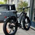 Zwarte fatbike e-bike met dikke banden en bagagedrager, geparkeerd voor een winkelpui.