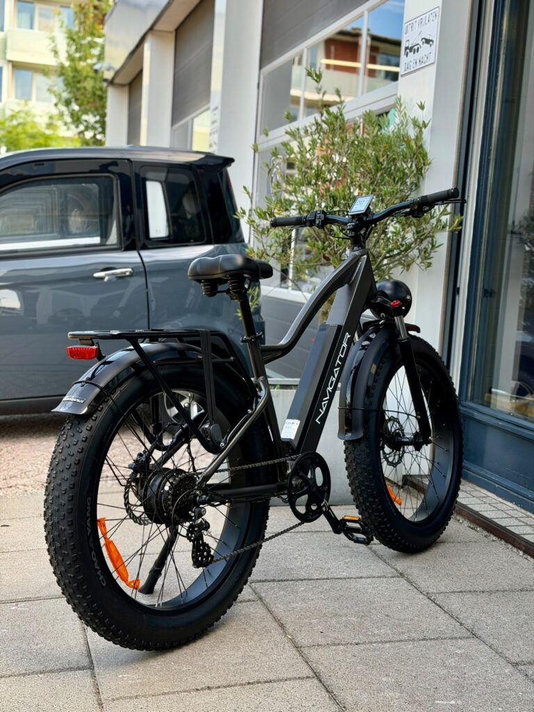Zwarte fatbike e-bike met dikke banden en bagagedrager, geparkeerd voor een winkelpui.