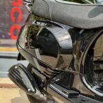 Close-up van een glanzende zwarte scooter met uitlaat en zadel, geparkeerd op straat.