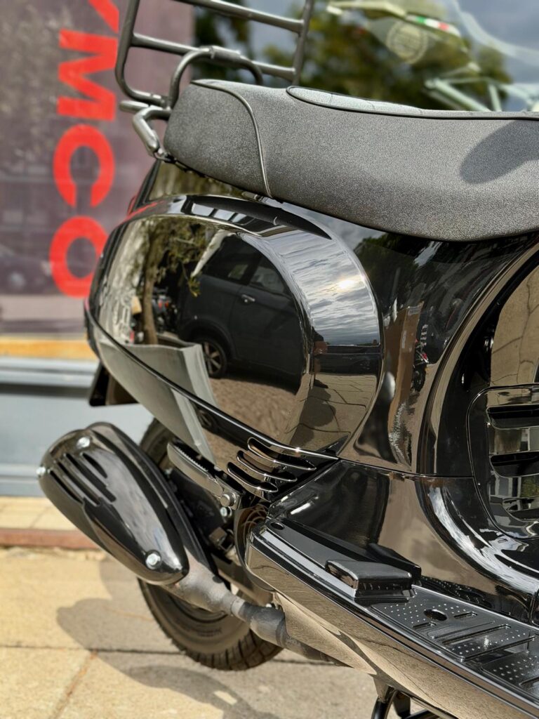 Close-up van een glanzende zwarte scooter met uitlaat en zadel, geparkeerd op straat.