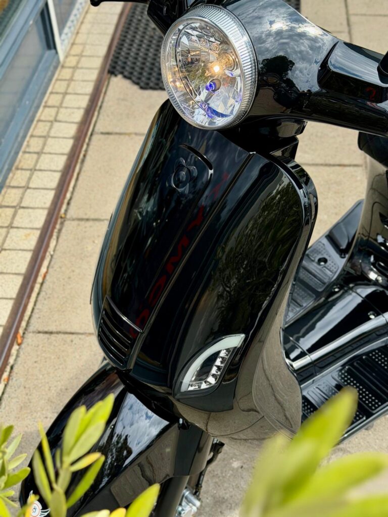 Close-up van de voorkant van een glanzende zwarte scooter met ronde koplamp en LED-dagrijlichten.