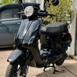 Zwarte scooter met ronde koplamp en bagagerek, geparkeerd op stoep bij garage.